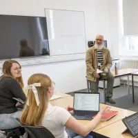 Alt text: A teacher sits on a desk holding papers while three students face them, one using a laptop. A whiteboard behind the teacher displays the phrase "POINT OF VIEW."